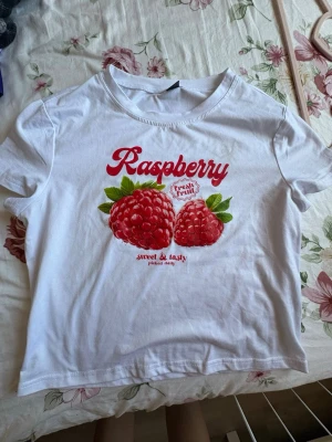 Vit t-shirt med hallontryck - Säljer en vit t-shirt med ett stort färgglatt hallontryck och texten 'Raspberry' i rött framtill. T-shirten har rund hals och korta ärmar. Perfekt för dig som gillar fruktiga och lekfulla prints.