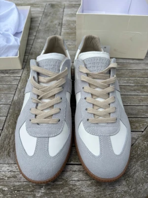 Vita och grå sneakers med mockadetaljer - Snygga Maison margiela gats i vitt och ljusgrått med beige snören och gummisula. Ovandelen är i en mix av slätt läder och mocka, vilket ger en stilren och modern look. Perfekta för dig som gillar minimalistisk design och neutrala färger.