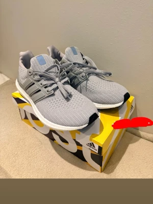 Grå Adidas Ultra Boost sneakers - Säljer ett par grå Adidas Ultra Boost sneakers med stickad ovandel och klassiska tre ränder på sidan. Skorna har vita Boost-sulor och grå detaljer på häl och snörning. Perfekta för dig som gillar stilrena och bekväma sneakers.