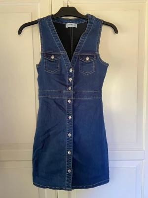Ärmlös jeansklänning med knappar - Snygg ärmlös jeansklänning i blå denim med v-ringning och knappar hela vägen framtill. Två bröstfickor med lock och kontrastsömmar ger en cool retrovibe. Perfekt till sommaren och lätt att styla med t-shirt eller tröja under.