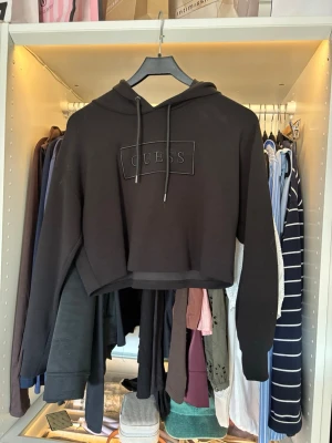Svart cropped hoodie från Guess - Snygg svart cropped hoodie från Guess med broderad logga på bröstet. Typ träningsmaterial