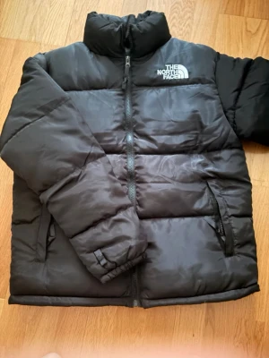  The North Face - Säljer en svart pufferjacka från The North Face med hög krage och dragkedja framtill. Jackan har två sidofickor med dragkedja och logga på bröstet. Perfekt för kalla vinterdagar och riktigt skön att bära.