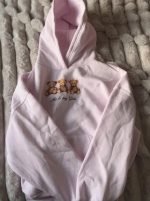 Ljusrosa hoodie med nalletryck - Söt ljusrosa hoodie med huva och magficka. På bröstet finns ett tryck med tre nallar och texten 'Me & my Gals'. Perfekt för dig som gillar gulliga detaljer och en avslappnad stil.