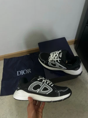 Svarta Dior sneakers  - Snygga svarta Dior sneakers med storlek 43 Helt ny och aldrig använt. Skorna har en sportig siluett med stora CD-loggor på sidan och vita skosnören. Materialet är en mix av textil och läder för en modern look.