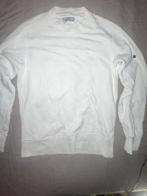 Vit sweatshirt från Stone Island - Klassisk vit sweatshirt från Stone Island med rund halsringning och ribbade muddar. Tröjan har en enkel och clean design med diskret logga på ärmen. Perfekt till en avslappnad och stilren look.