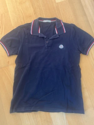 Mörkblå pikétröja från Moncler - Snygg mörkblå pikétröja från Moncler med klassisk krage och knappar framtill. Tröjan har Moncler-logga på bröstet och röda, vita och blå detaljer på krage och ärmslut. Tillverkad i bomull för en bekväm känsla.