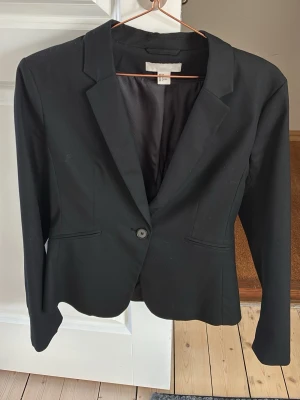 Svart klassisk kavaj med knapp - Stilren svart kavaj med en knapp framtill och två fickor. Kavajen har figurnära passform, klassisk krage och helfodrad insida. Perfekt för en snygg och uppklädd look. Materialet känns mjukt och följsamt.