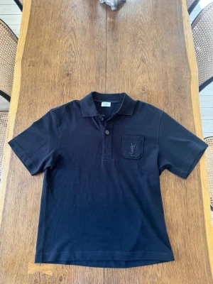 Svart pikétopp från Saint Laurent - Klassisk svart pikétopp från Saint Laurent med korta ärmar och krage. Toppen har knappslå framtill och en bröstficka med broderad YSL-logga. Perfekt för en stilren och avslappnad look.