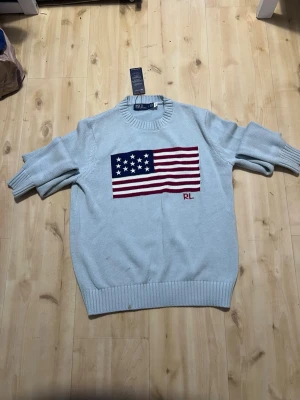 Ljusblå stickad tröja Polo Ralph Lauren - Ljusblå stickad tröja från Polo Ralph Lauren med amerikansk flagga broderad på bröstet och RL-detalj. Tröjan har rund halsringning och ribbade muddar vid ärmslut och nederkant. Perfekt för dig som gillar klassisk och stilren design.