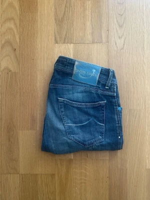 Mörkblå jeans från Jacob Cohen - Snygga mörkblå jeans från Jacob Cohen med klassisk femficksmodell och raka ben. Jeansen har en diskret tvättad look och patch med logga bak i midjan. Perfekta till en avslappnad stil och tillverkade i mjukt denimtyg. Det finns ett litet hål vid skrevet som är igensytt i samma färg som jeansen så det är inget som syns.