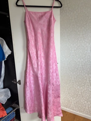 Lång rosa klänning med blommönster - Säljer en lång, ärmlös klänning i ljusrosa med blommigt mönster. Klänningen har smala axelband och är tillverkad i ett glansigt material som ger en elegant känsla. Perfekt för sommarens alla tillfällen!