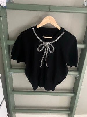 Svart topp med strassrosett - Snygg svart topp med korta ärmar och rund halsringning. Dekorerad med en stor rosett i strass framtill som ger en lyxig och trendig känsla. Perfekt för dig som vill sticka ut lite extra. Materialet känns mjukt och stretchigt.