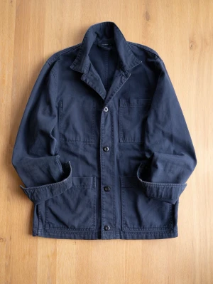 Navy Chore Jacket, size M - Stilren mörkblå overshirt med klassisk krage och tre stora utanpåfickor. Jackan har knäppning framtill med svarta knappar och är tillverkad i ett slitstarkt bomullstyg. Perfekt för lager på lager och enkel att matcha med olika outfits.