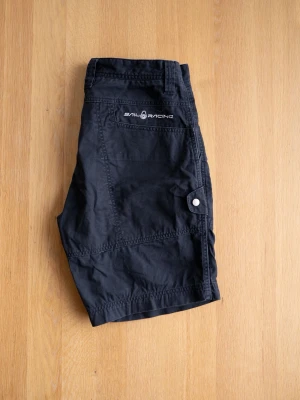 Svarta shorts från Sail Racing, size S - Svarta shorts från Sail Racing med flera fickor, bälteshällor och knappstängning. Tillverkade i bomull för en bekväm känsla. Perfekta för varma dagar och har en stilren look med diskret logga på ena fickan.