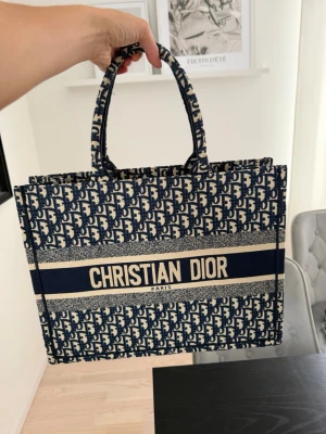 Tote handväska - Snygg Book Tote handväska. Väskan har två handtag och rymligt inre fack, perfekt för att bära med sig allt man behöver. Klassisk och trendig design med tydlig logga framtill.