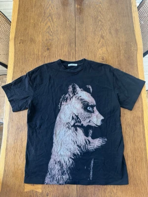 Svart Acne t-shirt  - Cool svart acne t-shirt med ett stort tryck av en björn på framsidan. T-shirten har rund hals och korta ärmar, perfekt för en avslappnad stil. Materialet känns mjukt och bekvämt, troligen bomull.