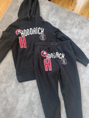 Svart hoodrich set - Svarta mjukisbyxor från Hoodrich med stora broderade detaljer och loggor i vitt och rött. Byxorna har dragsko i midjan och matchande hoodie finns. Perfekta för en avslappnad streetwear-look.