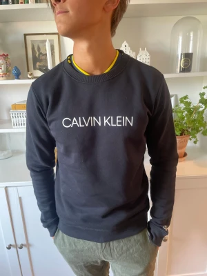 Svart sweatshirt från Calvin Klein - Svart sweatshirt från Calvin Klein med vit logga tryckt på bröstet. Tröjan har rund halsringning, långa ärmar och ribbade muddar vid ärmslut och nederkant. Perfekt för en avslappnad och stilren look.
