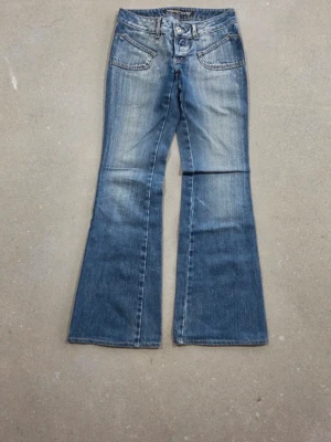 Blå lågmidjade bootcut jeans  - Säljer ett par blå bootcut jeans från Salt River med snygg slitning och breda ben. Jeansen har två framfickor med dekorativa sömmar och klassisk knappgylf.