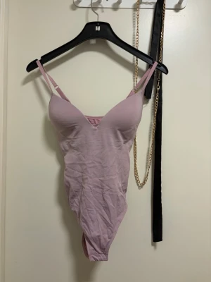 Ljuslila/rosa body med smala axelband - Säljer en ljuslila body med smala axelband och vadderade kupor. Bodyn har en djup urringning och är tillverkad i ett mjukt, stretchigt material som formar sig efter kroppen. Perfekt för att skapa en snygg siluett under kläder.