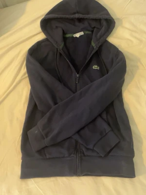 Mörkblå hoodie från Lacoste - Snygg mörkblå hoodie från Lacoste med dragkedja framtill och klassisk krokodillogga på bröstet. Modellen har huva och ribbade muddar vid ärmslut och nederkant. Perfekt för en chill och sportig look.