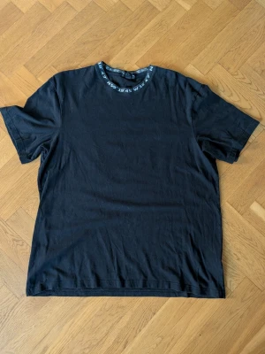 Svart t-shirt med tryck på kragen - En svart t-shirt i bomull med korta ärmar och rund hals. Kragen har ett tryck med siffror och bokstäver som ger en cool detalj. Perfekt till jeans eller shorts för en avslappnad stil.