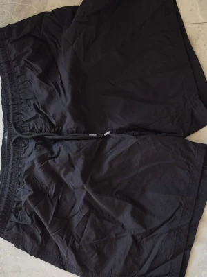 Svarta shorts med snörning XXL - Svarta shorts med elastisk midja och snörning. Shortsen har innerfoder i mesh för extra komfort och andningsförmåga. Perfekta för varma dagar eller träning. Enkel och stilren design utan synliga loggor eller mönster.