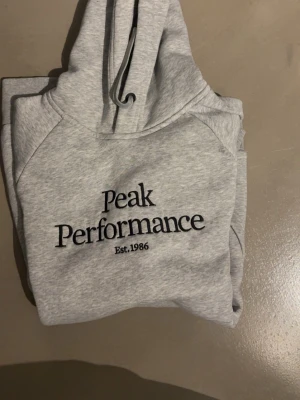 Grå hoodie Peak Performance  - Snygg ljusgrå hoodie från Peak Performance med svart broderad logga på bröstet. Klassisk huva med snörning och mjukt material i bomull och polyester. Perfekt för en avslappnad och sportig stil.