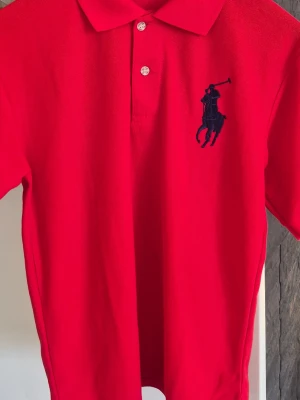 Röd pikétröja med broderad logga - Snygg röd pikétröja med klassisk krage och knappar framtill. Stor broderad logga i marinblått på bröstet och en broderad siffra 9 på ärmen. Tillverkad i bomull för en bekväm känsla. Perfekt till jeans eller shorts.