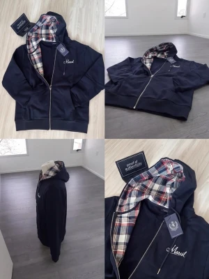 Marcel marinblå hoodie med rutigt foder - Snygg marinblå hoodie från Marcel med dragkedja och huva. Insidan av huvan har ett rutigt foder i rött, vitt och svart som ger en unik touch. Broderad logga på bröstet. Perfekt för dig som vill ha en stilren men ändå personlig hoodie.