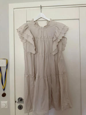 Beige volangklänning från Object - Söt beige kortklänning från Object med volangdetaljer på axlarna och hög krage. Klänningen har en luftig passform och är tillverkad i ett lätt bomullstyg. Perfekt för varma dagar och ger en romantisk vibe.