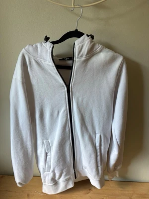 Vit hoodie med dragkedja och huva - Säljer en enkel och stilren vit hoodie med dragkedja framtill och två fickor. Hoodien har justerbar huva med snören och ribbade muddar vid ärmar och nederkant. Perfekt för en avslappnad look och passar till det mesta.