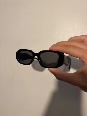 Prada solglasögon  - Snygga svarta solglasögon från Prada med rektangulära, kantiga bågar och mörka glas. Tjocka skalmar med Prada-logga på sidan ger en lyxig och trendig vibe. Perfekta för dig som vill sticka ut med en statement-accessoar.