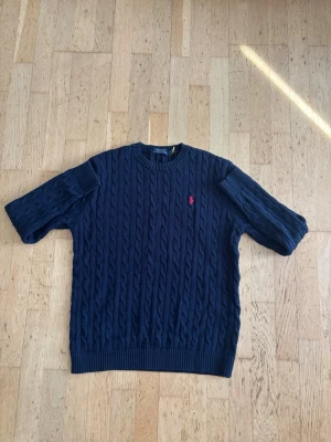 Polo Ralph lauren kabelstickad tröja  - Kabelstickad mörkblå tröja från Polo Ralph Lauren med rund halsringning och ribbade muddar. Tröjan har det klassiska röda Polo-logot broderat på bröstet och är tillverkad i mjukt material. Perfekt för en stilren och avslappnad look.