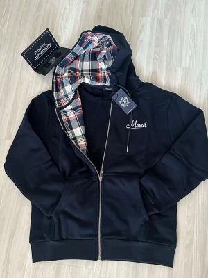 Svart hoodie med rutigt foder - Snygg svart hoodie med dragkedja och huva. Insidan av huvan har ett rutigt foder i rött, vitt och blått. På bröstet finns en liten vit brodyr med texten 'Marcel'. Perfekt för en avslappnad och trendig stil.
