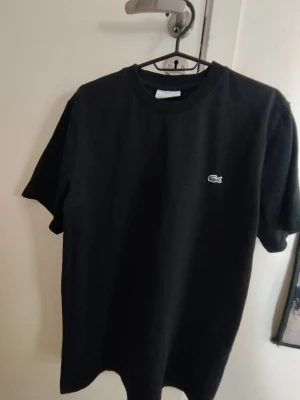 Lacoste T-shirt - Klassisk svart t-shirt från Lacoste med den ikoniska krokodilloggan på bröstet. T-shirten har rund hals och korta ärmar, perfekt för en clean och stilren look. Tillverkad i mjuk bomull för skön komfort.