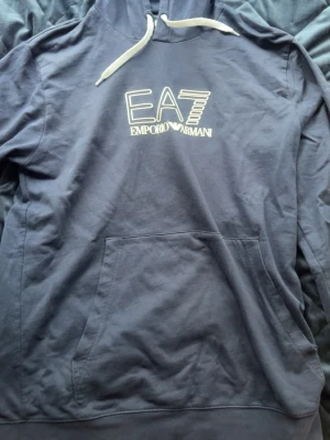 Mörkblå hoodie EA7 Emporio Armani - Snygg mörkblå hoodie från EA7 Emporio Armani med vit logga på bröstet och vita dragsnören i huvan. Klassisk känguruficka framtill och mjukt material som känns skönt mot huden. Perfekt för en avslappnad och trendig look.