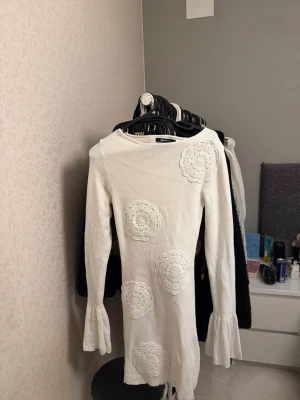 Vit stickad klänning med spetsdetaljer - Säljer en vit stickad klänning med långa ärmar och volang vid ärmsluten. Klänningen har dekorativa virkade spetsapplikationer framtill som ger en unik look. Modellen är kroppsnära och kort, perfekt för dig som gillar en feminin och trendig stil. Den sitter utmärkt och formar sog efter din kroppsform😍råkade beställa två utav samma och deog av prislappen så vill bli av med den!
