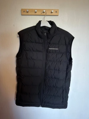 Svart dunväst Peak Performance - Snygg svart dunväst från Peak Performance med hög krage och dragkedja framtill. Västen har diskret logga på bröstet och quiltad design som ger en sportig look. Perfekt att slänga över en hoodie eller tröja för extra värme.