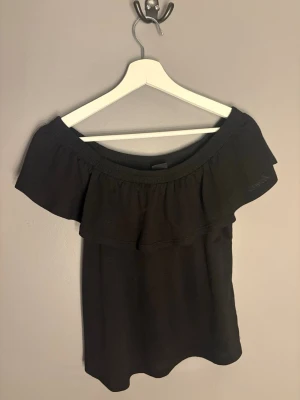 Svart offshoulder topp med volang - Snygg svart offshoulder topp med bred volang runt axlarna. Perfekt för dig som vill ha en trendig och enkel topp till sommaren. Tillverkad i mjukt material och har en avslappnad passform.