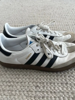 Adidas Samba vita sneakers - Klassiska Adidas Samba sneakers i vitt läder med svarta detaljer och bruna gummisulor. Skorna har de ikoniska tre ränderna på sidan och texten 'SAMBA' i guld. Låg modell med snörning och mockadetaljer vid tån. Perfekta för en sportig och stilren look.