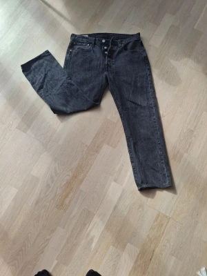 Levi's 501 Jeans - Klassiska Levi's 501 raka jeans med knappgylf, svart/antracit-tvätt. Etiketten säger W31 L30 men de är uppklippta till L27 innerbenslängd — mät gärna mot ett par egna jeans innan köp. Äkta läderlapp på baksidan, röd flik, alla original Levi Strauss & Co-knappar intakta. Bra skick — inga hål eller fläckar, bara ärligt slitage. Tillverkade i USA.