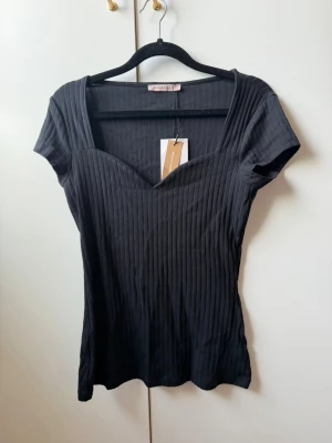 Svart ribbad topp från Anna Field - Snygg svart ribbad topp från Anna Field med v-ringad hals och korta ärmar. Toppen har en figurnära passform och är tillverkad i mjuk bomullsmix. Perfekt att styla med jeans eller kjol för en trendig look.