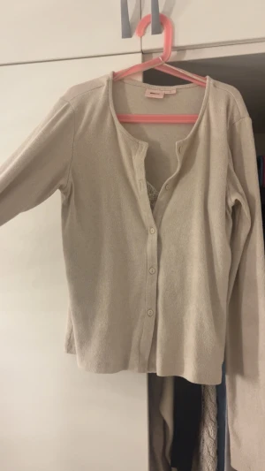Beige ribbad kofta med knappar - Stilren beige kofta i ribbat material med långa ärmar och knäppning framtill. Koftan har en enkel, rak passform och insytt spetslinne.