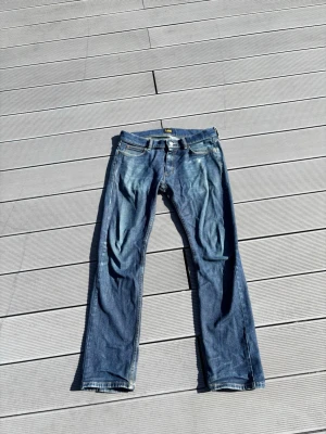 Blå Lee slim fit jeans - Snygga blå slim fit jeans från Lee med klassisk femficksdesign och slitningar på låren. Jeansen har orange kontrastsömmar och metallknapp med Lee-logga. Perfekta för dig som gillar en avslappnad men ändå stilren look.