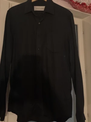Svart långärmad skjorta - Stilren svart skjorta med klassisk krage och långa ärmar. Skjortan har knappar framtill och en enkel bröstficka. Perfekt för dig som gillar en clean och tidlös look.
