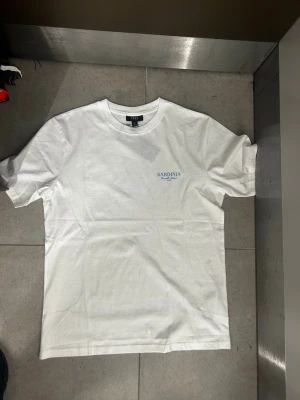 Vit t-shirt med Sardinia-tryck - Vit t-shirt med tryck fram och stort färgglatt motiv på ryggen med texten 'La Dolce Vita Sardinia'. T-shirten har rund hals och korta ärmar. Materialet ser ut att vara mjuk bomull, perfekt för en chill och stilren look. Finns i många olika storlekar 