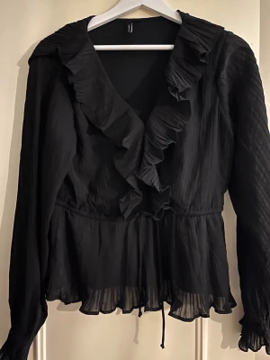 Svart blus med volang och knytband från Veromoda - Stilren svart blus med djup V-ringning och bred volangkant längs urringningen. Blusen har långa ärmar och ett knytband i midjan för en feminin siluett. Materialet är lätt och något transparent med plisserad struktur. Perfekt för en trendig look.