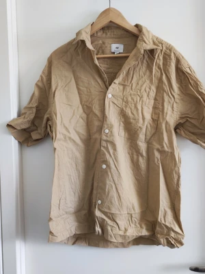 Beige linneskjorta från H&M - Säljer en beige skjorta från H&M i relaxed fit. Skjortan är tillverkad i 100% linne, har korta ärmar, klassisk krage, bröstficka och vita knappar framtill. Perfekt för en avslappnad och stilren look.