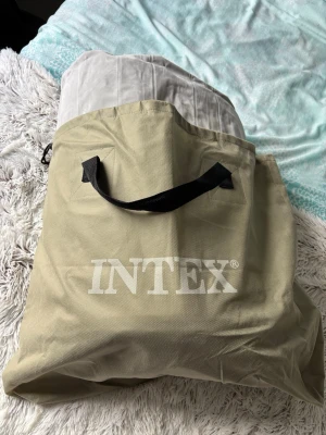 Luftmadrass Intex med förvaringsväska - Säljer en grå luftmadrass från Intex som kommer med en beige förvaringsväska med svart handtag. Madrassen är stor och har en mjuk ovansida i ljusgrått och mörkgrå kanter. Perfekt för gäster eller camping.
